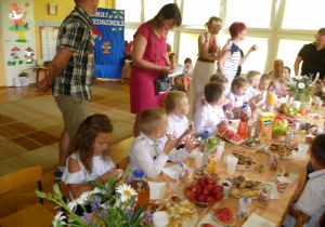 Dzieci siedzą za stołami zastawionymi słodkim poczęstunkiem.