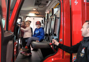Adaś, Laura i Oliwia siedzą w samochodzie strażackim.