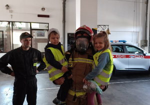 Julka i Radek na rękach strażaka w pełnym stroju do akcji ratowniczej.