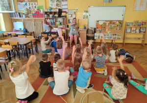 Dzieci siedzą na dywanie ,unoszą ręce i łączą je ze sobą.
