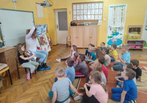Dzieci siedzą na dywanie. Mama Neli pokazuje ilustrację w książce.