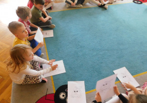 Dzieci siedzą na dywanie trzymając kolorowanki sowy.