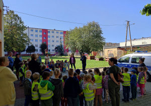 Dzieci stoją z Paniami i Panem Policjantem na placu w ogrodzie.