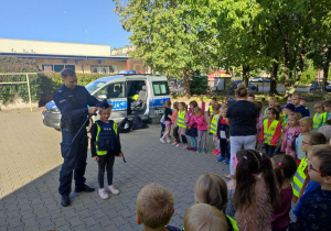 Karinka ubrana w czapkę i kamizelkę kuloodporną z Panem Policjantem stoją przed dziećmi.