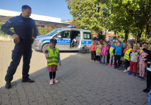 Dziewczynka stoi z Panem Policjantem przed dziećmi i pokazuje tarcze do kierowania ruchem.