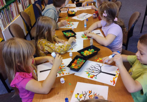 Dzieci siedzą przy stolikach i wyklejają papierem jesienne drzewa.