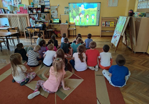 Dzieci siedzą na dywanie, oglądają film edukacyjny.
