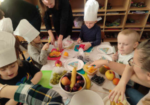 Dzieci z mamami przy stolikach przygotowują owocowe i warzywne przekąski.