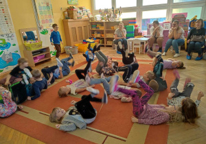 Dzieci leżą na plecach na dywanie i ruszają nogami.