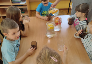 Dzieci siedzą przy stolikach i piją wodę smakową.