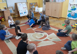 Dzieci siedzą na dywanie i słuchają opowieści czytanej przez nauczycielkę.