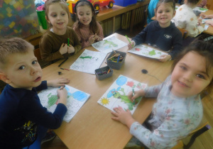 Dzieci siedzą przy stoliku i kolrują obrazek Dinka na łące.