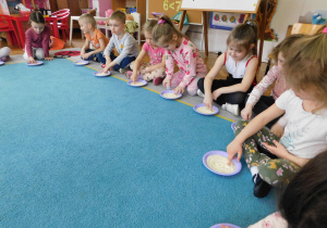 Dzieci siedzą na dywanie i rysują w kaszy na talerzykach cyfrę "O".