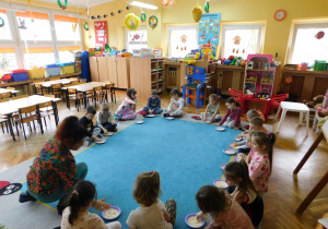 Dzieci z grupy "Biedronek" siedzą na dywanie i rysują w kaszy na talerzykach cyfry.