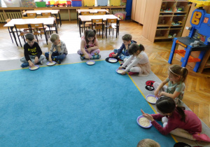Dzieci siedzą na dywanie i rysują w kaszy na talerzykach cyfrę "3".