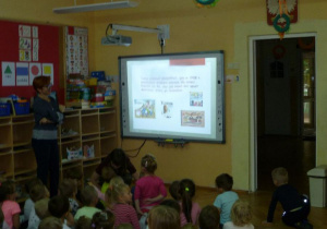  Prowadząca czyta tekst i pokazuje obrazy na ekranie, grupa dzieci siedzi i ogląda.