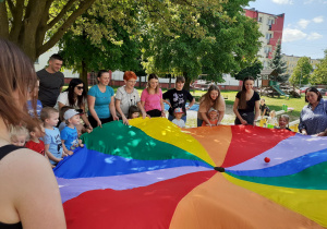 Rodzice i dzieci trzymając chustę próbują trafić piłeczką do otworu po środku.