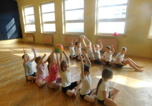 Dzieci siedzą w dwóch rzędach i podają sobie piłki nad głowami.