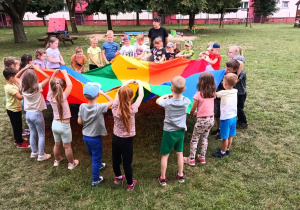 5 Dzieci stoją na trawie w kole z chustę animacyjną, równomierne ją podnosząc i opuszczając.