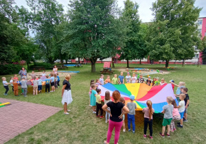 Przedszkolaki trzymają chustę i naprowadzają małą piłeczkę, tak, żeby wpadła do otworu w środku chusty.