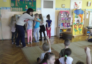  Sześcioro dzieci stoi odwróconych tyłem do grupy siedzącej na dywanie, prowadząca wskazuje jedno ze zgłaszających się dzieci do odpowiedzi;