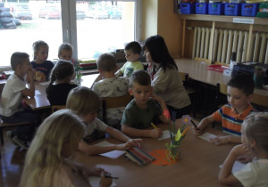 Dzieci rysują kredkami przy stolikach na małych kartkach; prowadząca kuca między dziećmi i wyjaśnia zadanie;