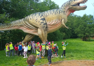Przedszkolaki stoją pod ogromną figurą dinozaura.