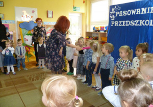 Dzieci stoją w półkolu, Pani Dyrektor dotyka ramienia dziecka dużą kredką, pasując na przedszkolaka.